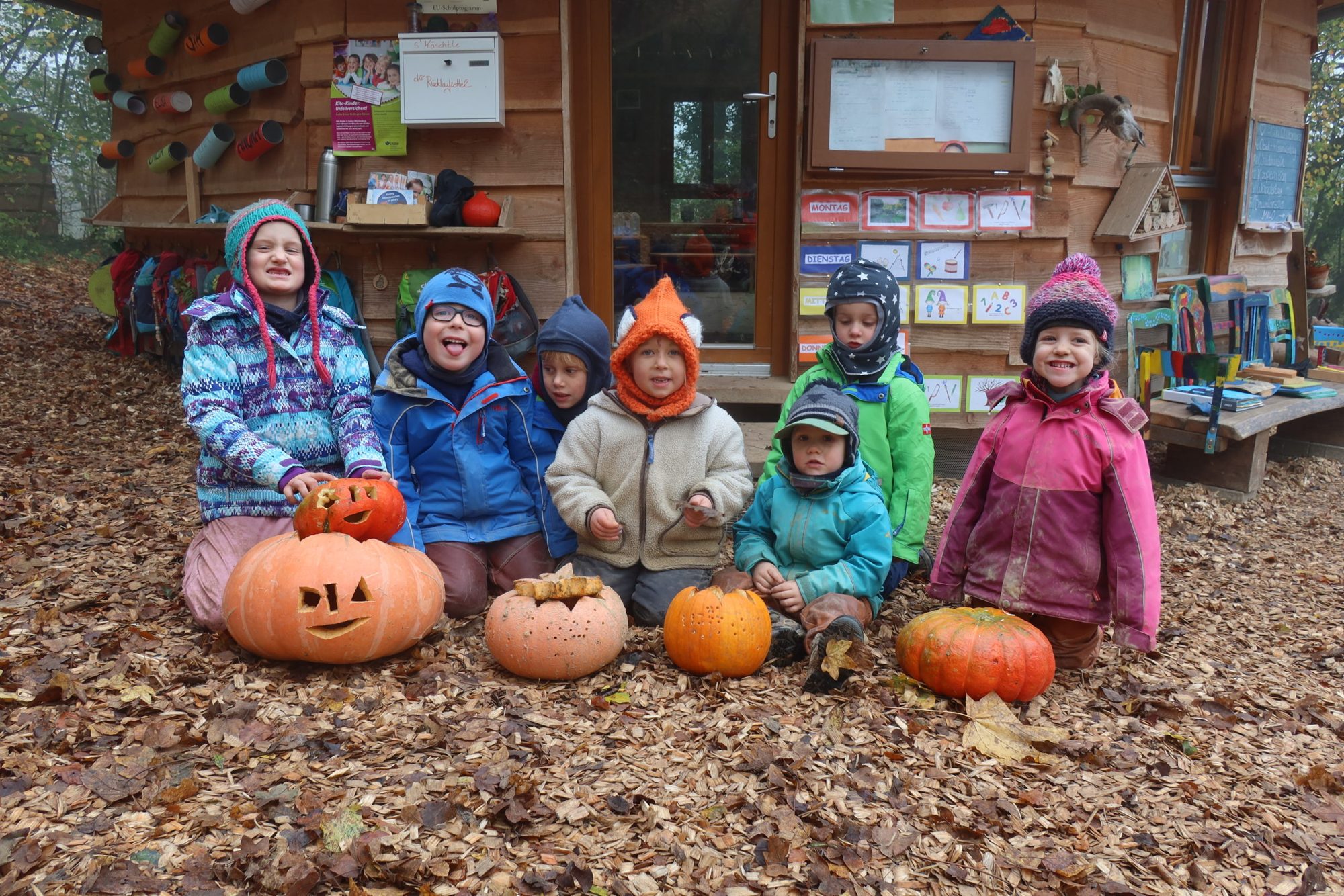 Kinder in Herbstkleidung mit geschnitzten Kürbissen vor der Holzhütte im Laub
