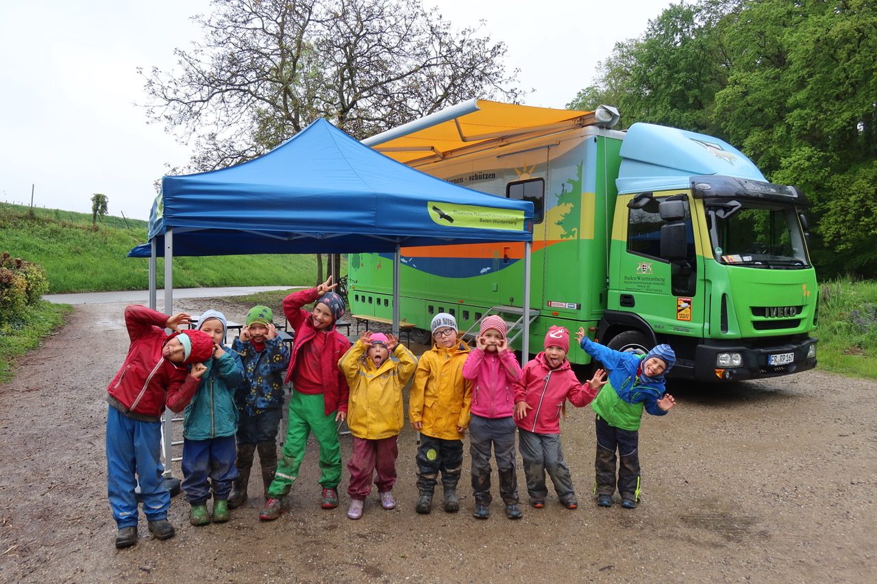 Kinder in Regenkleidung vor einem Pavillon und grünem Umweltmobil-Lastwagen