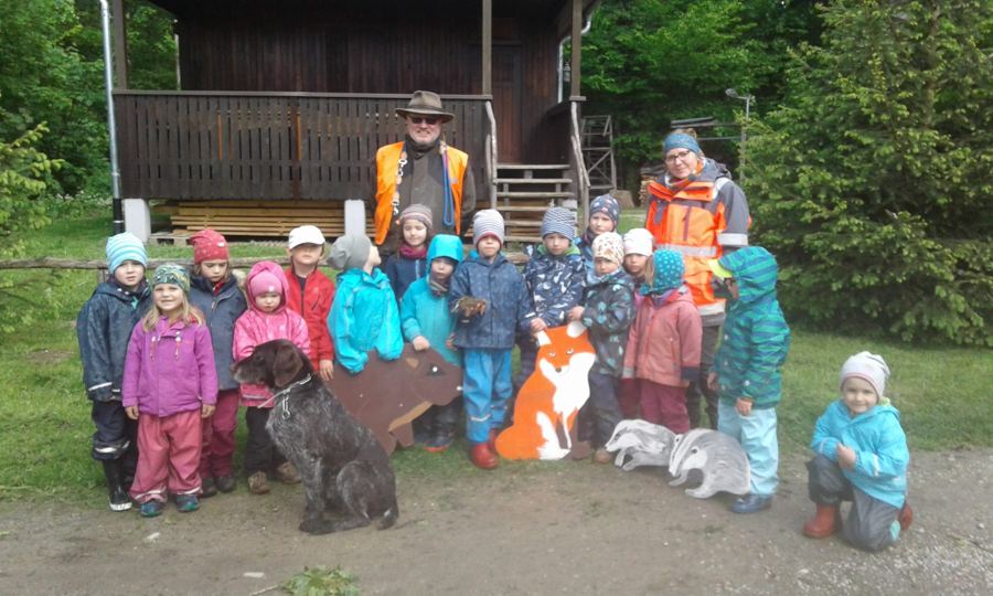 Kindergruppe mit Jäger, Erzieherin, Hund und Tierfiguren aus Holz vor einer Hütte