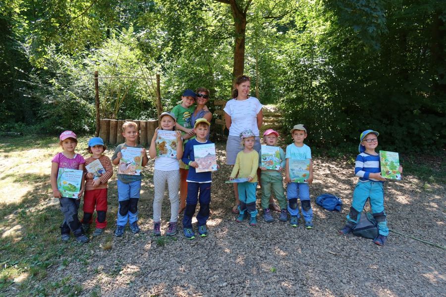 Kinder zeigen im Wald ihre Bücher, dahinter zwei Erzieherinnen und ein Kind